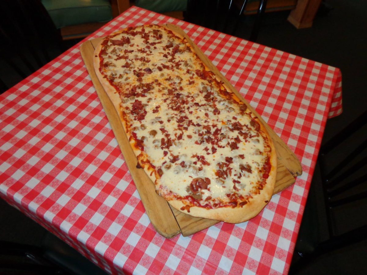 A thirty-inch football-shaped pizza on a wooden paddle, on a red and white checked tablecloth